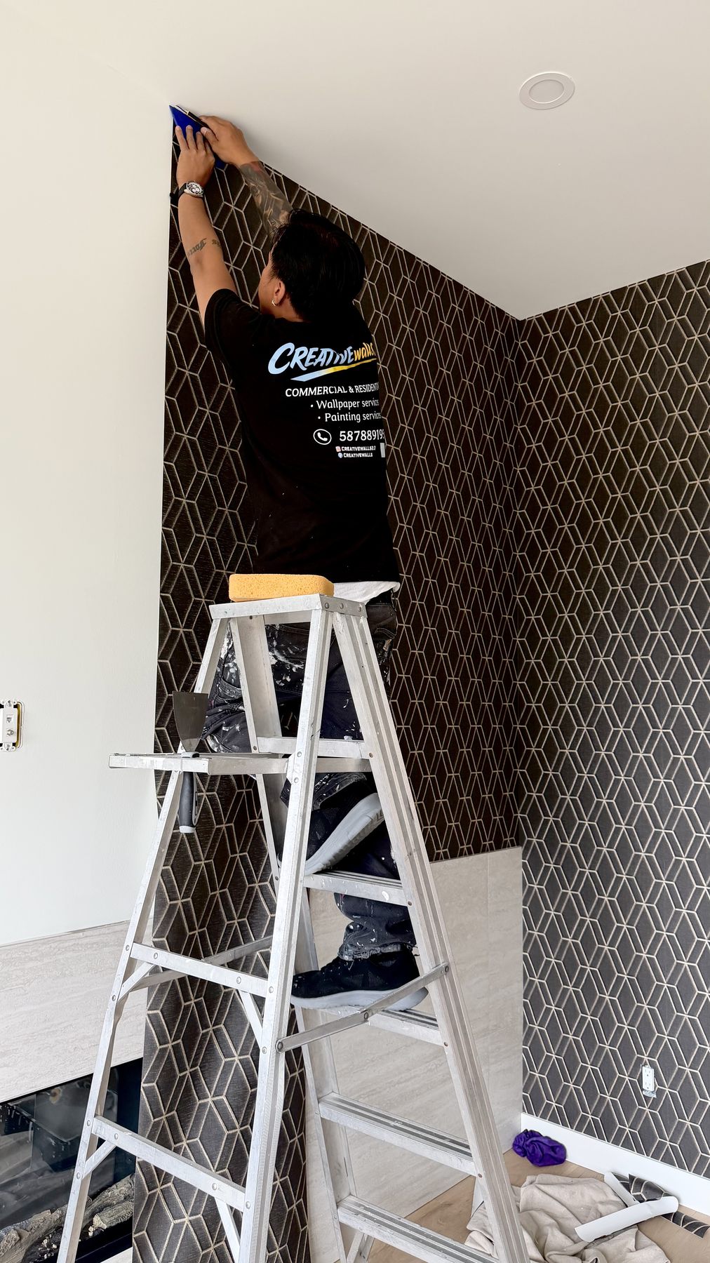 Installer in a Creative Walls shirt on a ladder, smoothing geometric wallpaper into a ceiling corner