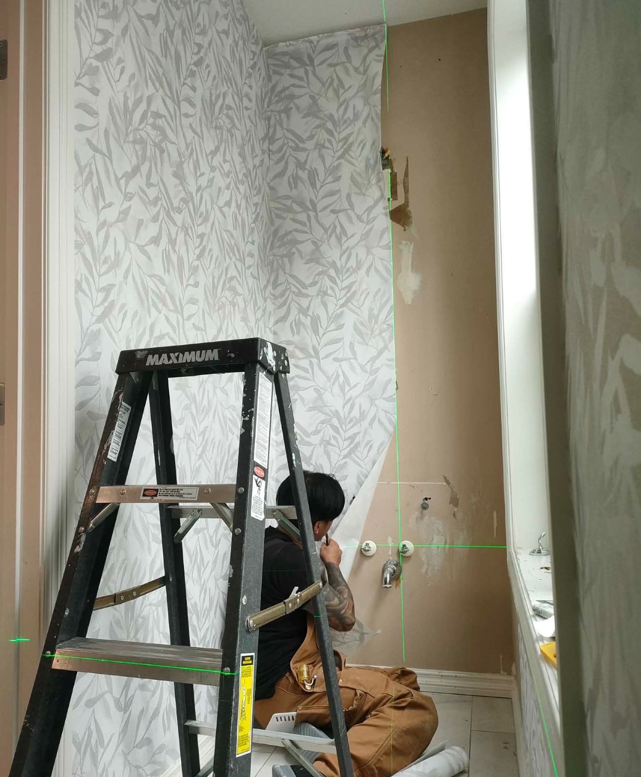 Installer kneeling beside a ladder, hanging leaf-pattern wallpaper in a bathroom with laser guide lines